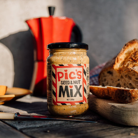 A jar of Pic's Seed & Nut Mix  styled on a rustic wooden table with sliced artisanal bread, a red moka pot, and a knife in a warm, natural light setting.